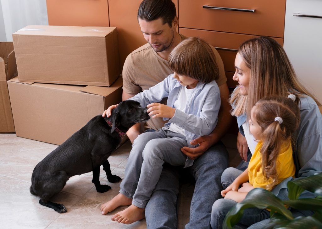 family of four, including two kids and a dog, sitting beside moving boxes