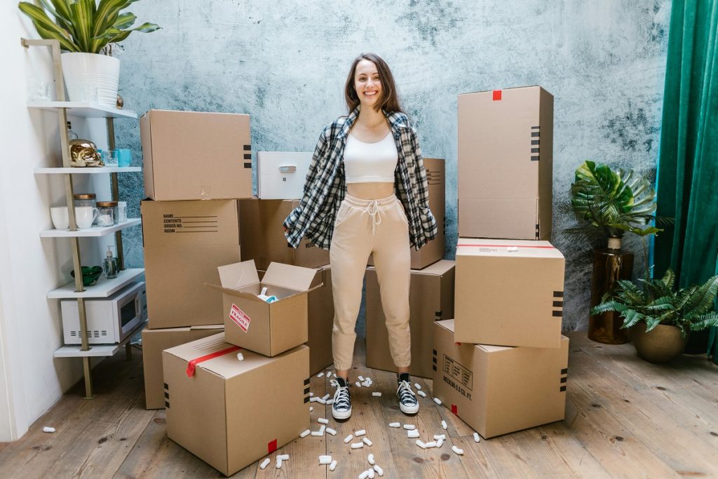 woman wearing a plaid shirt standing in a room surrounded by cardboard moving boxes and indoor plants