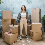 woman wearing a plaid shirt standing in a room surrounded by cardboard moving boxes and indoor plants